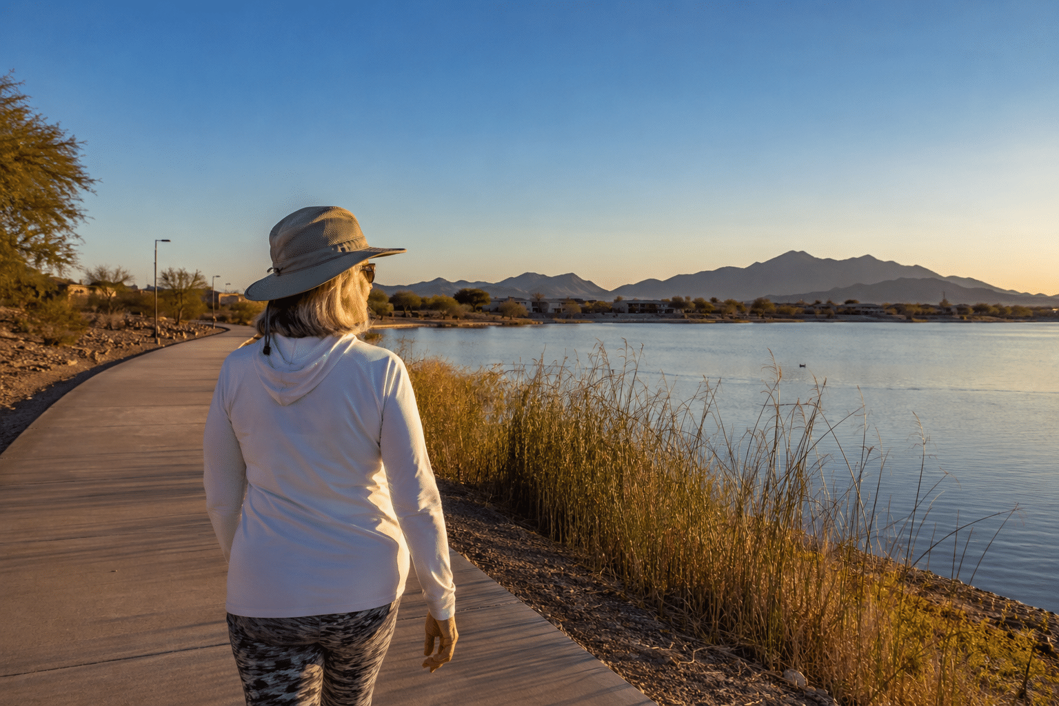 woman in her late 60s walking alone on a paved waterfront path along an engineered desert lake in Goodyear, Arizona